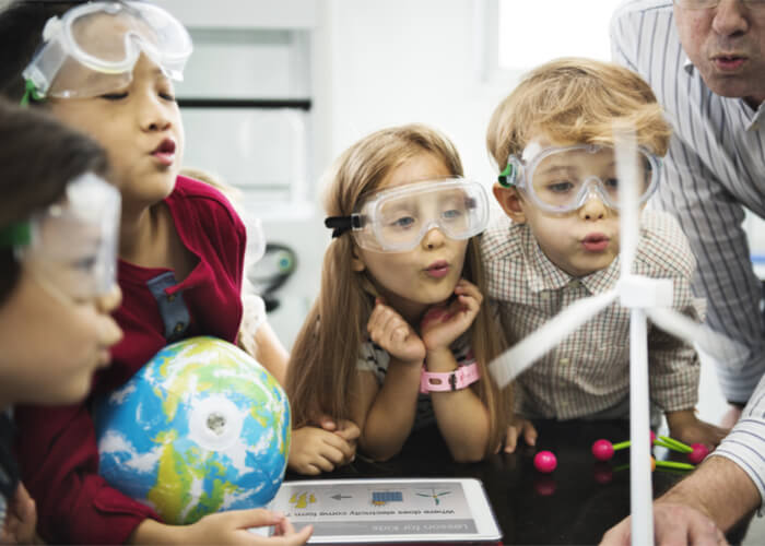 A photograph of children and a teacher learning about wind power, and blowing on a model of a wind turbine | Chaos Theory